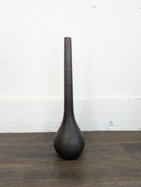 1. Tall, dark metal vase with a narrow neck and bulbous base, resting on a wooden floor against a white wall.