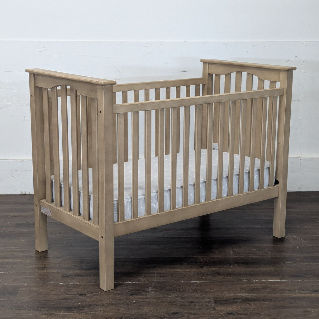 Angled side view of Pottery Barn Kids natural wood crib, slatted design, mattress included, on dark wood floor.