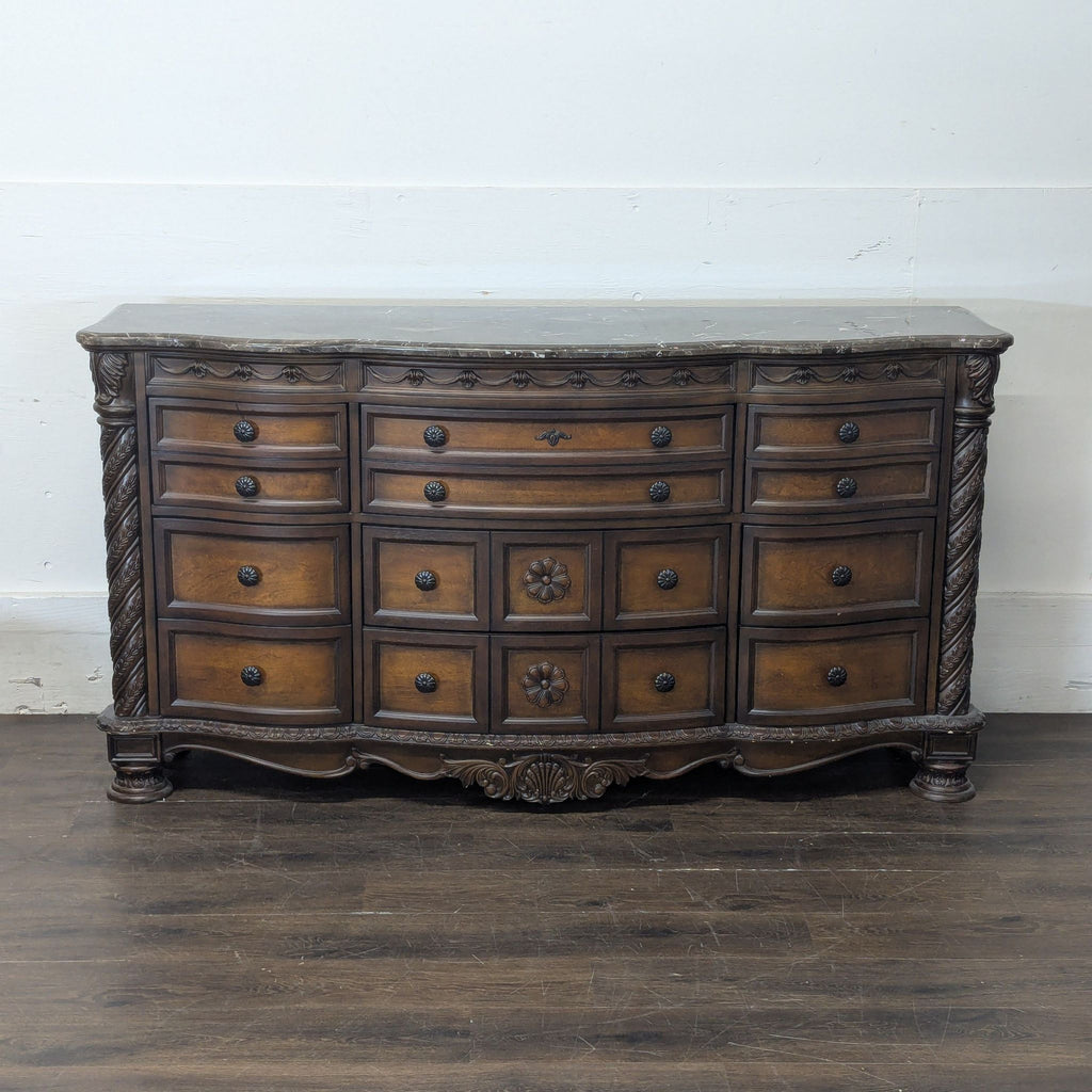 3. Formal Ashley Furniture dresser with a marble surface, detailed woodwork, and numerous drawers on hardwood floor.