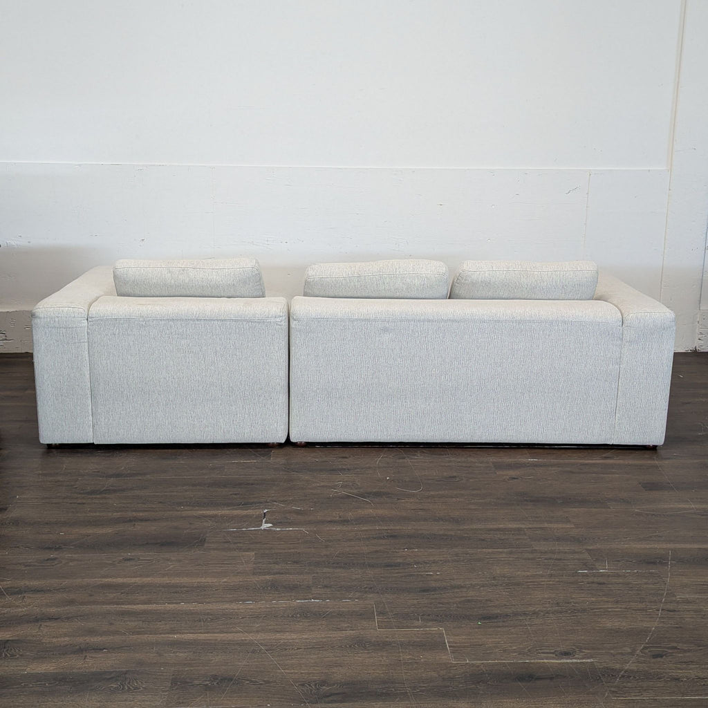 Back view of a light gray sectional sofa with three back cushions, set against a plain wall and wood floor.