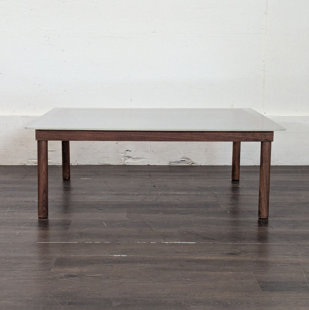 2. Minimalist Design Within Reach coffee table with a clear glass top and wooden frame, set against a plain white wall.