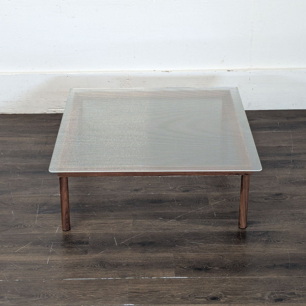 3. Square coffee table from Design Within Reach, featuring a ribbed glass top and simple wood legs, on dark flooring.