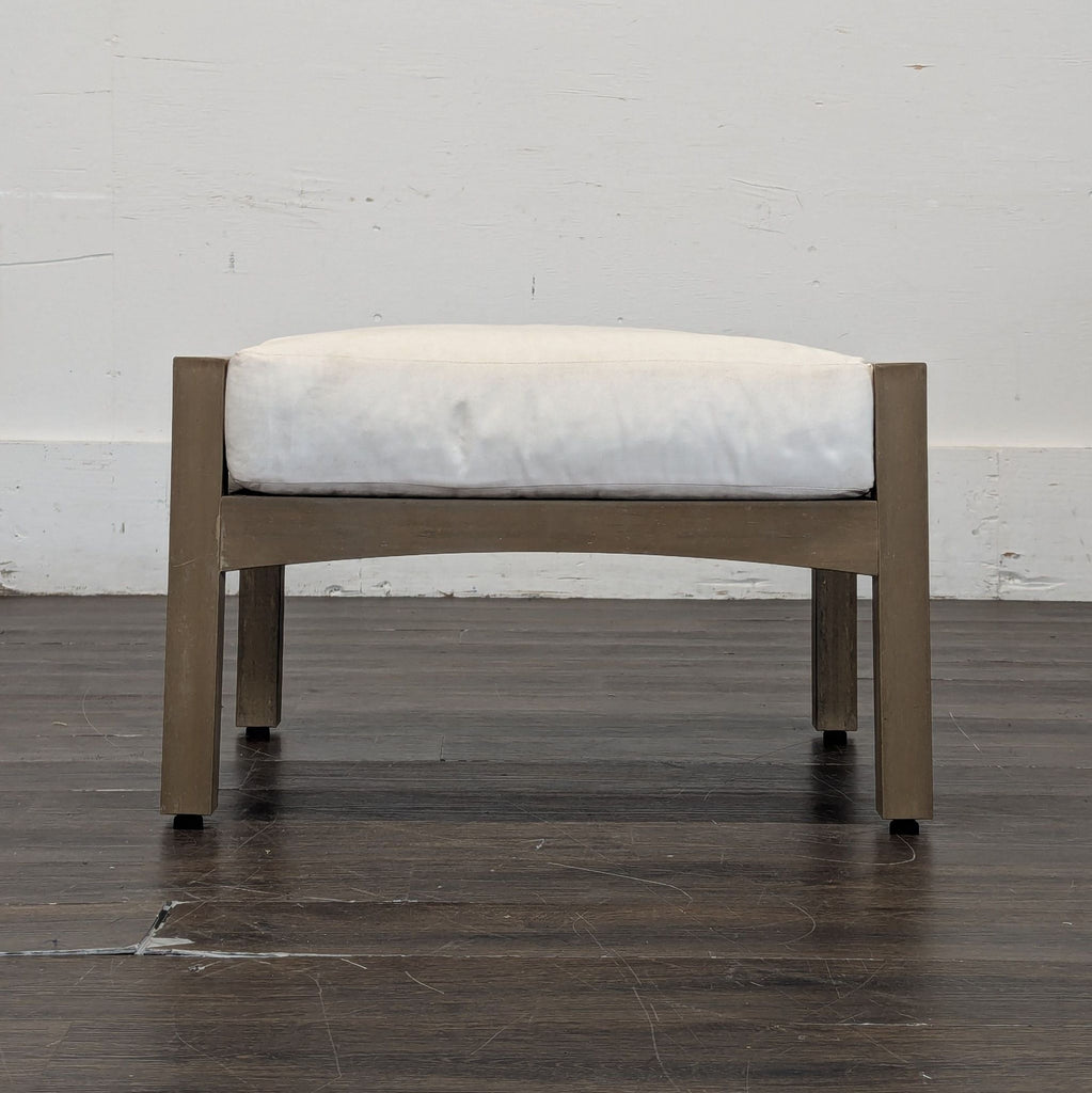 1. Square wooden ottoman with a light brown frame and white cushion, placed on a dark floor against a plain wall.  