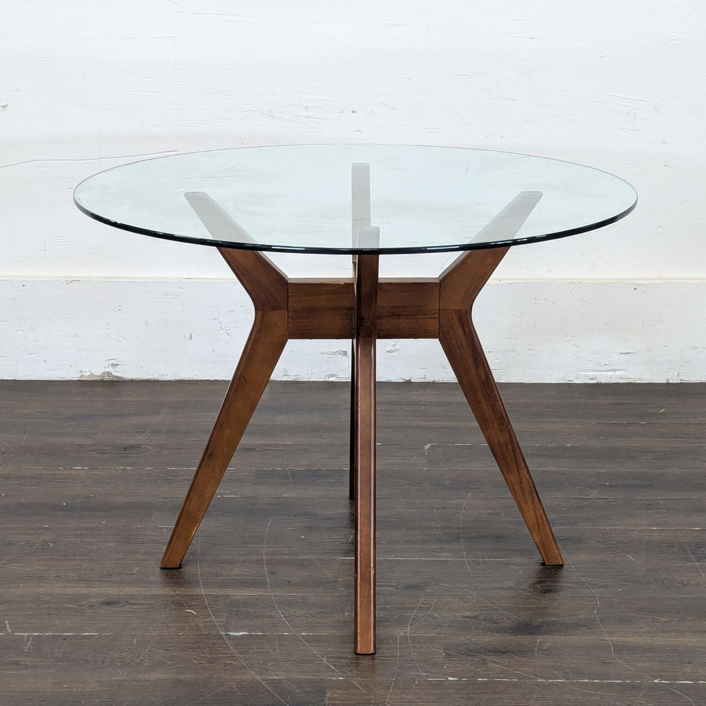 2. Glass round dining table with angled wooden legs, set against a plain wall and wood floor.