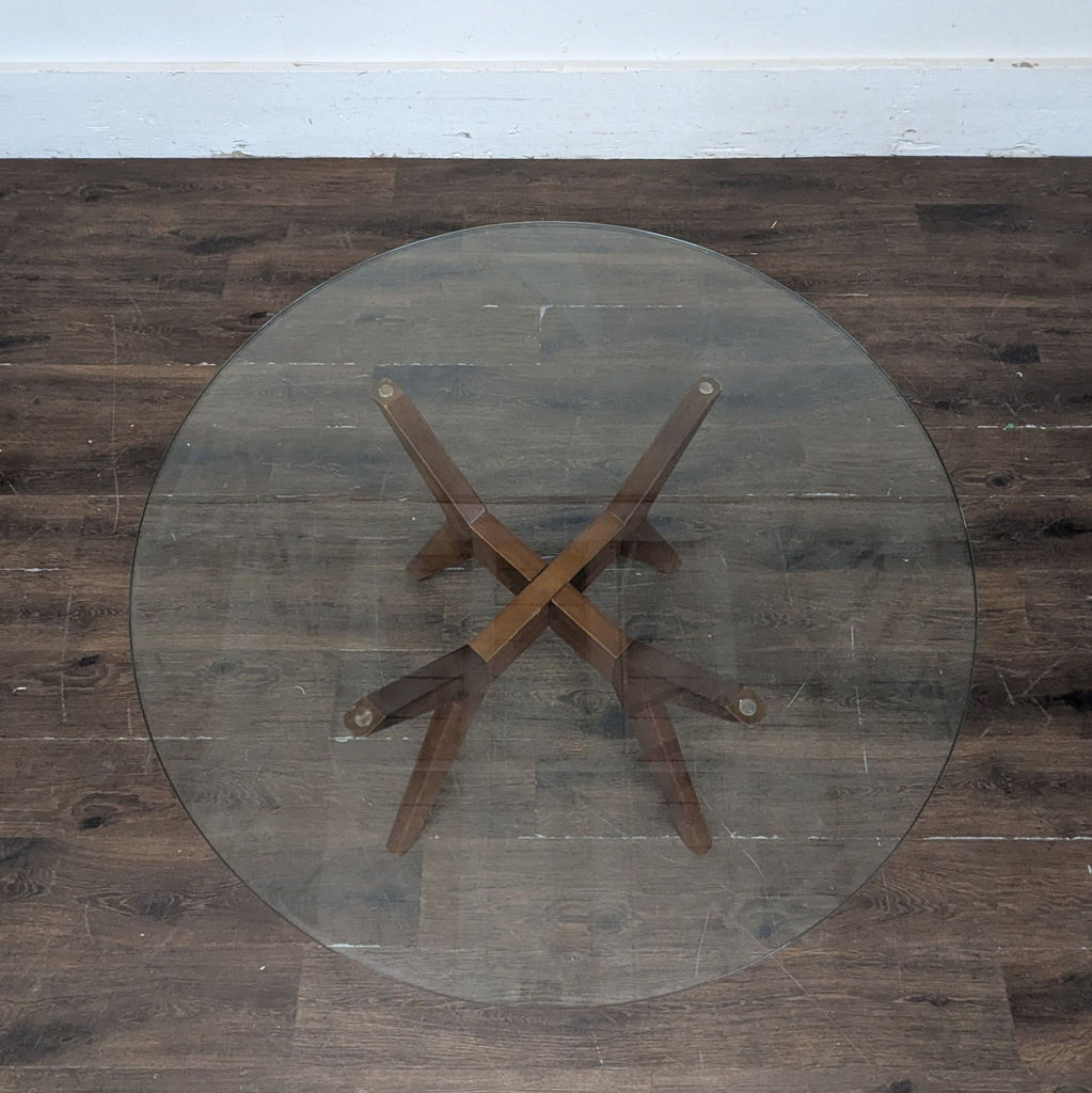 3. Top view of a round glass dining table with intersecting wooden legs on a wooden floor.
