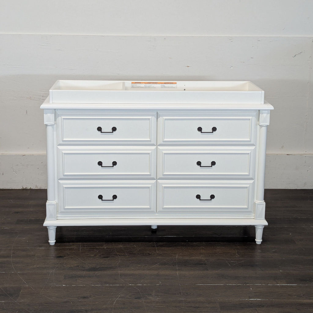 3. Simmons Juvenile white dresser with six drawers and black handles, placed against a plain wall on a wood floor.