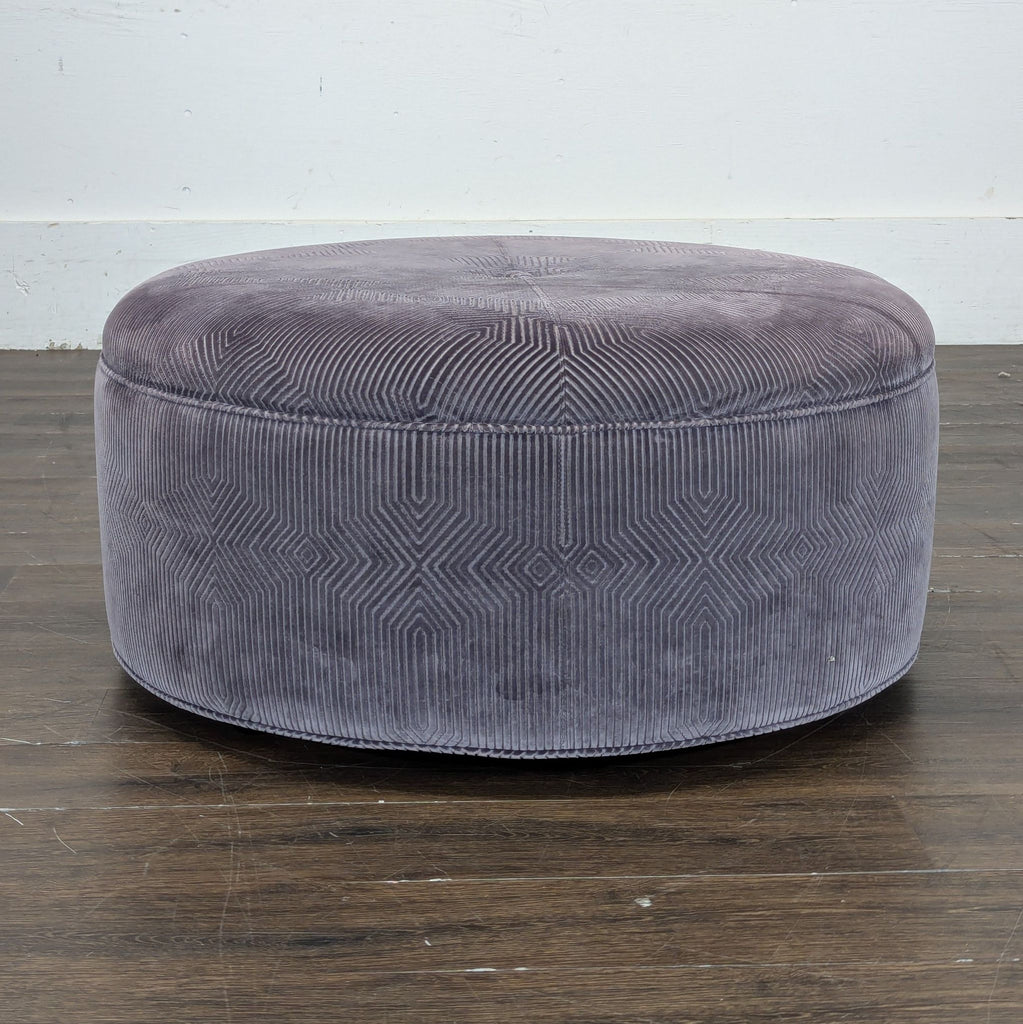 2. Side view of a circular Lexington ottoman in velvet with a modern geometric design, placed on dark wood flooring.