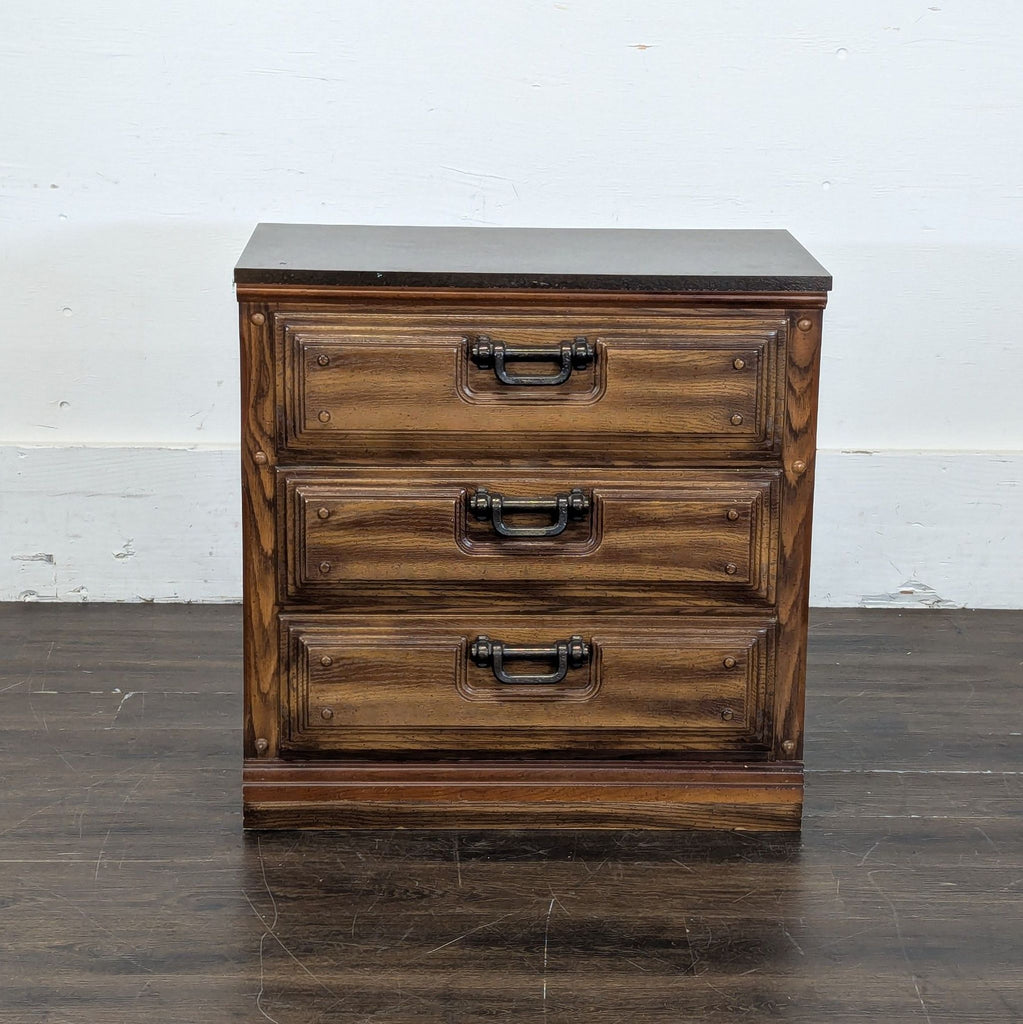 Front view of a small wooden dresser with three drawers and metal handles, set against a white wall.  