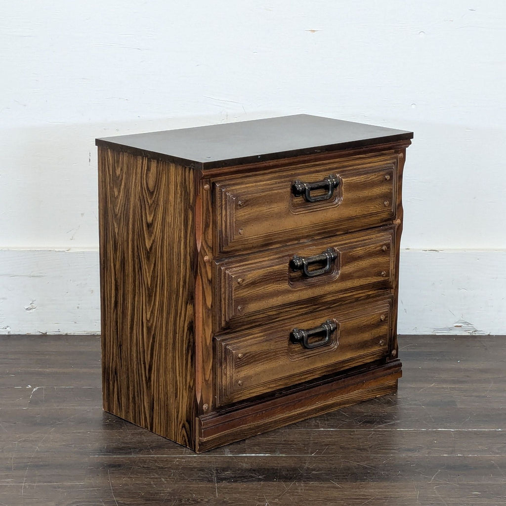 Angled side view of a three-drawer wooden dresser featuring dark metal handles and a flat top, on a wooden floor.