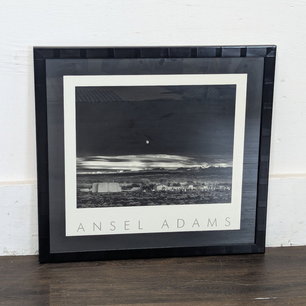 2. Framed Ansel Adams landscape print resting against a white wall on a wooden floor, clear and well-lit view.