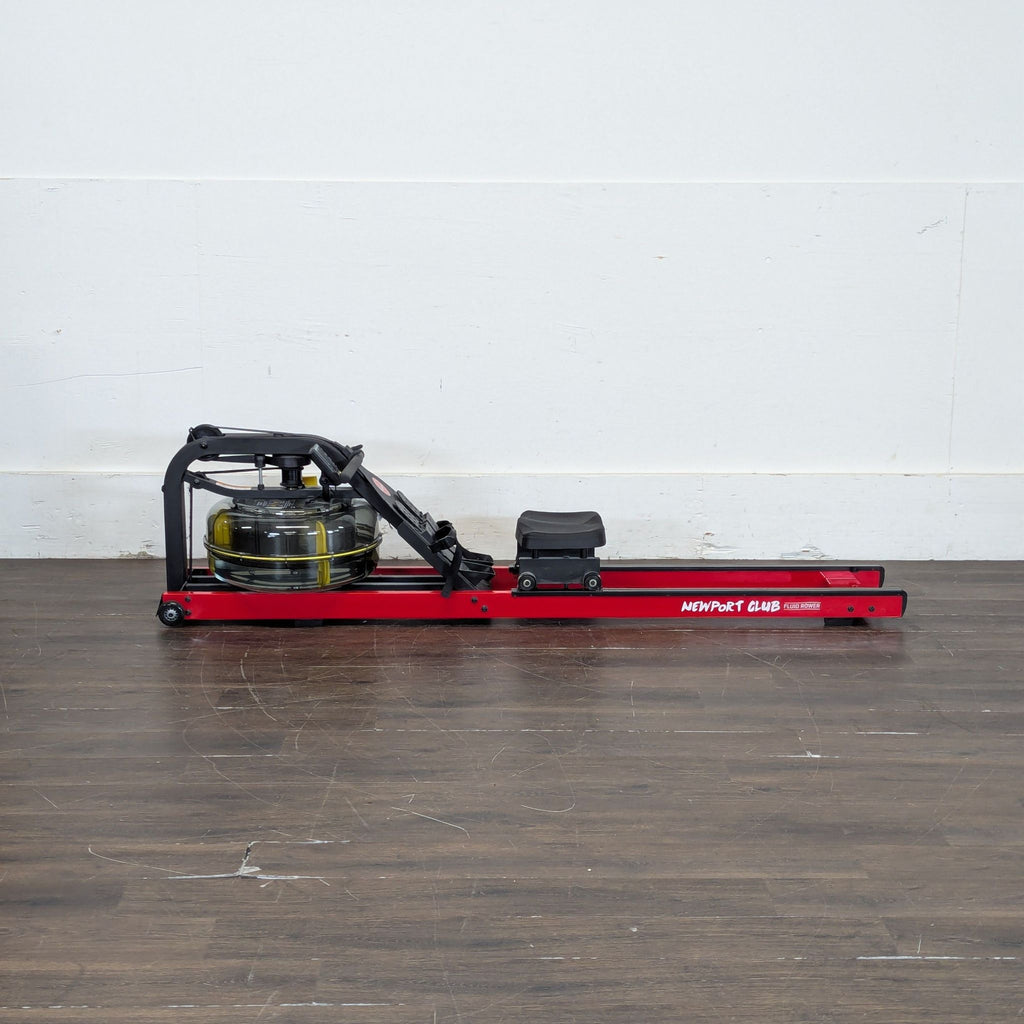 1. Side view of a First Degree Fitness rowing machine with a red frame and water resistance tank, placed on a wooden floor.