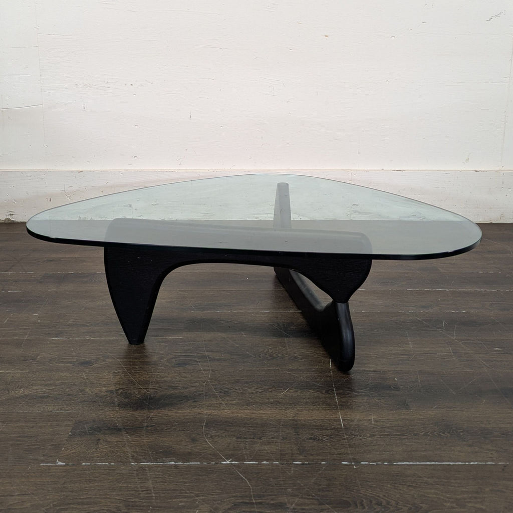 2. Modern coffee table with a clear glass top and sculpted black wooden base, shown on wooden flooring.