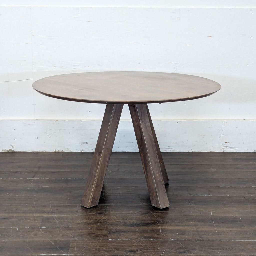 3. Simple round wood dining table with splayed legs, photographed against a plain wall on dark flooring.
