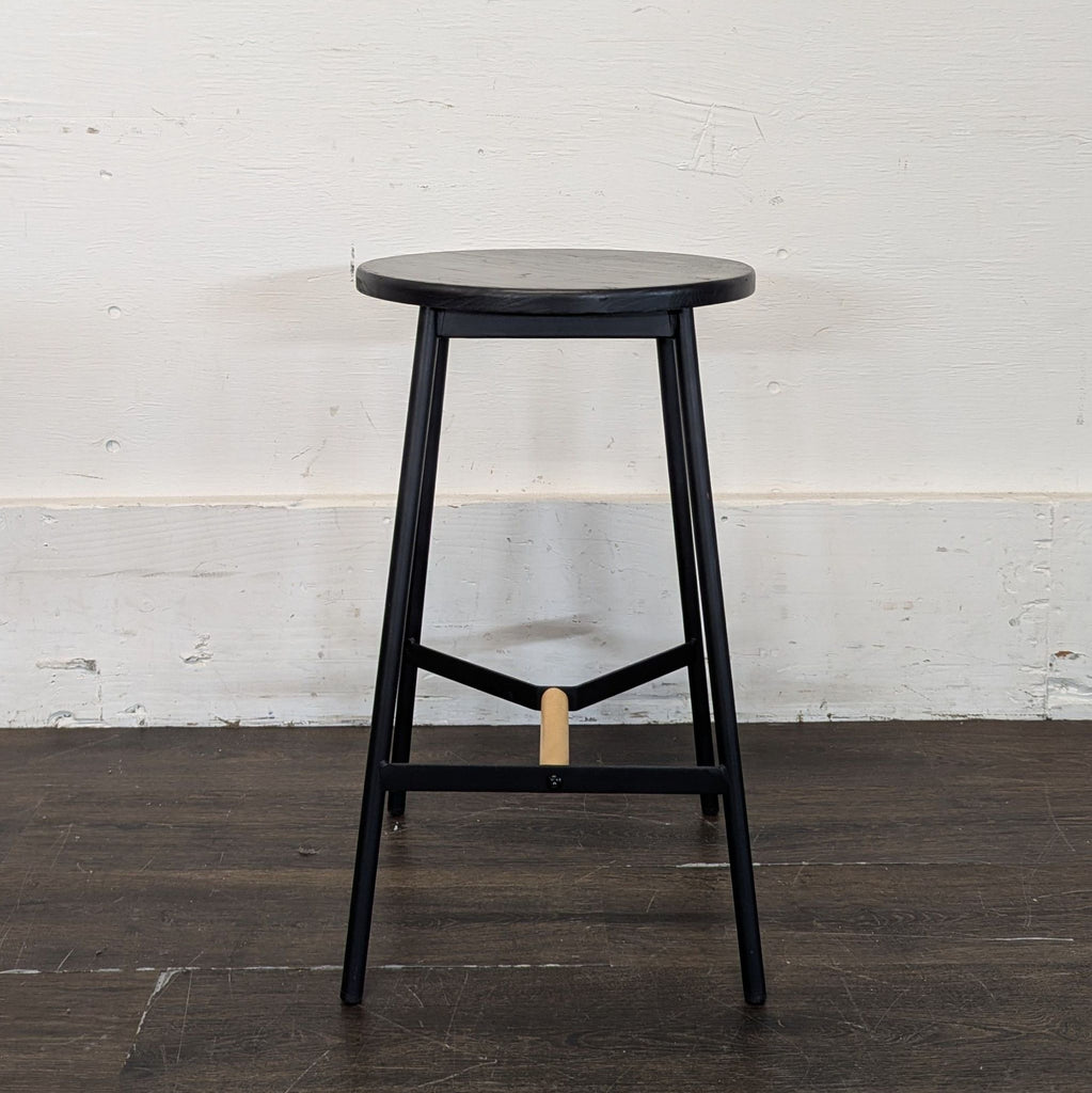 Single black stool with round seat, metal legs, and light wood footrest against a white wall.