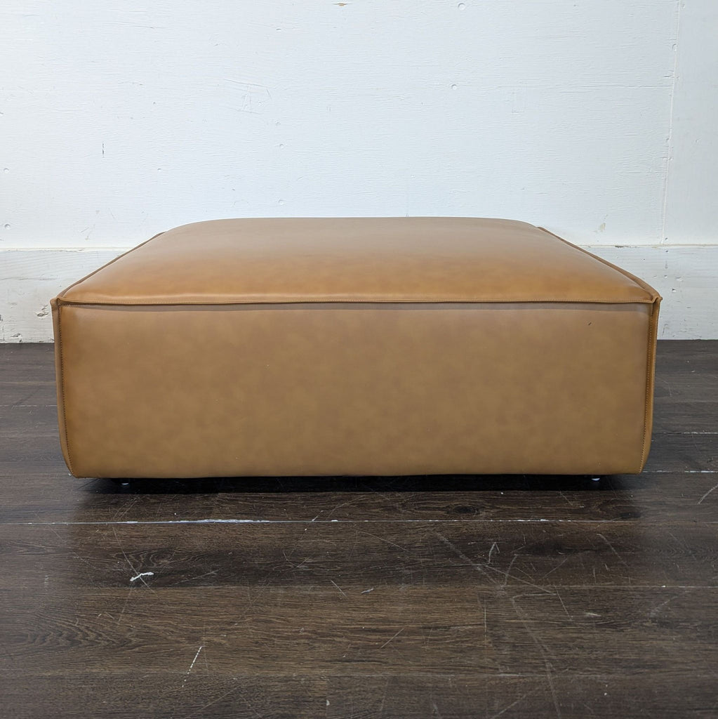 Low, wide, tan ottoman with leather-like finish shown from the front, placed on a wooden floor against a white wall.
