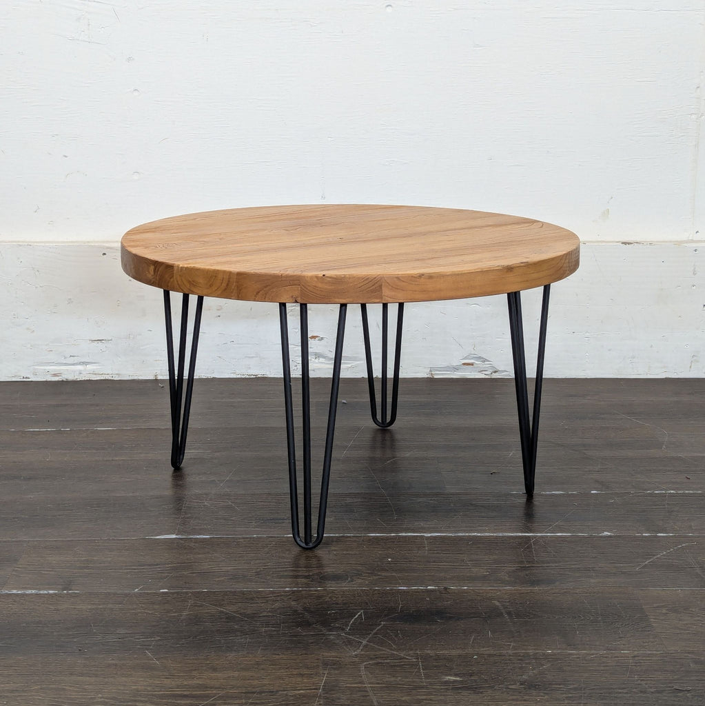3. Side angle of a round wooden coffee table with sleek black metal hairpin legs, set against a white wall background.