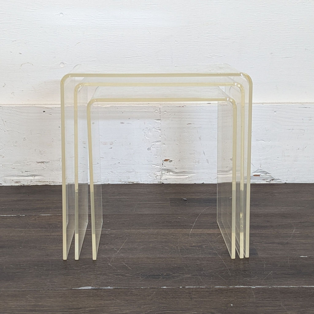 1. Set of two clear nested acrylic end tables with rounded corners on a dark wood floor against a white wall.