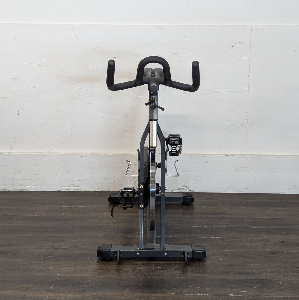 Front view of a BodyCraft stationary bike with black handlebars and pedals, positioned on a wooden floor.