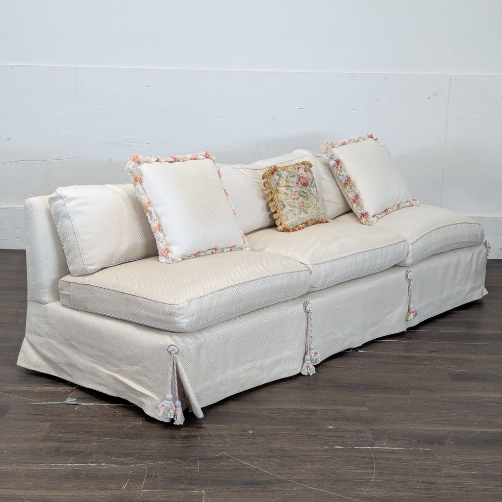 3. Side view of cream 3-seat sofa with tassel skirt and pastel-trim pillows, set against plain white wall.