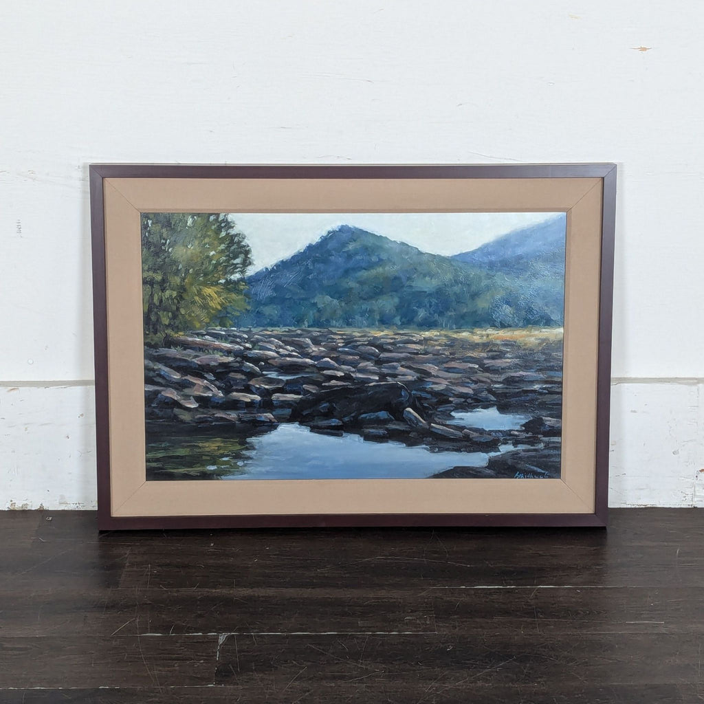 1. Framed painting of a rocky river scene with distant green hills and reflective water, set against a white wall.  