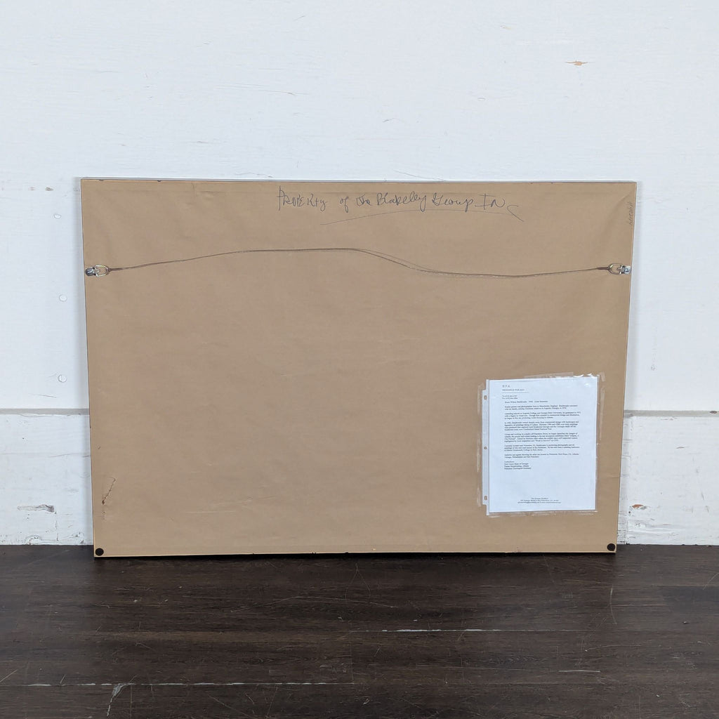 2. Back view of framed artwork with brown paper backing, hanging wire, and an attached document on the lower right.