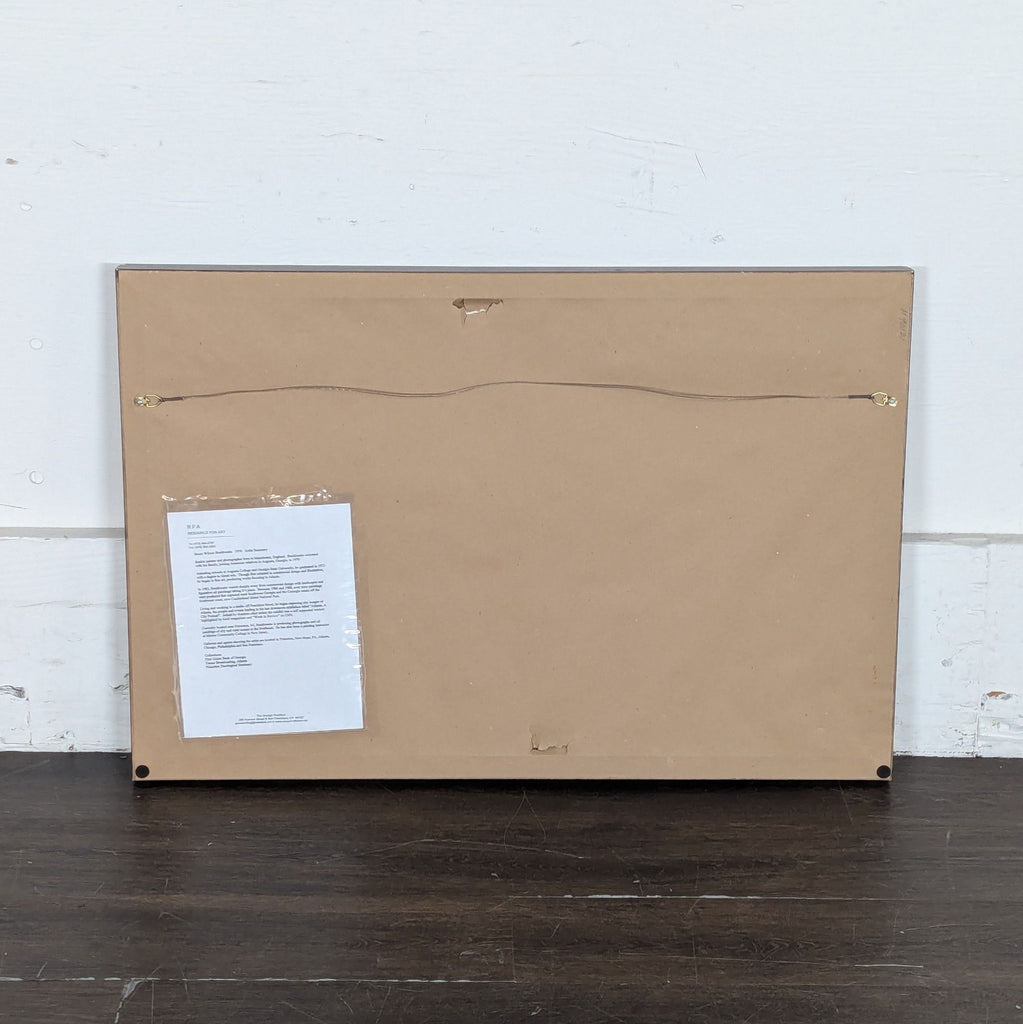 3. Back view of framed artwork with a wire hanger and an attached information sheet on the brown paper backing.