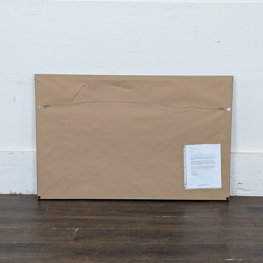 3. Back view of a framed artwork with brown paper backing, hanging wire, and a document attached to the lower right.