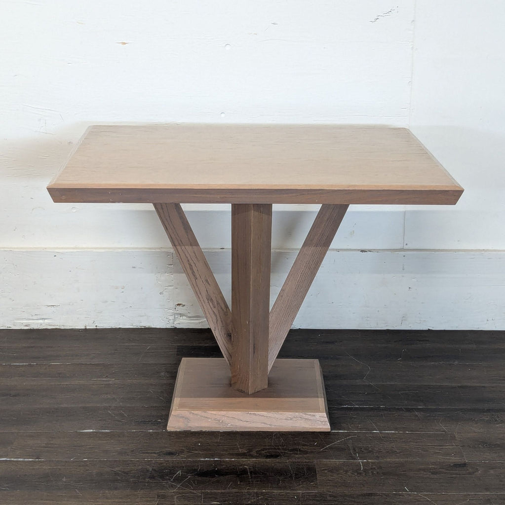 3. Wooden dining table with a simple square top and central V-base, positioned against a plain white wall indoors.