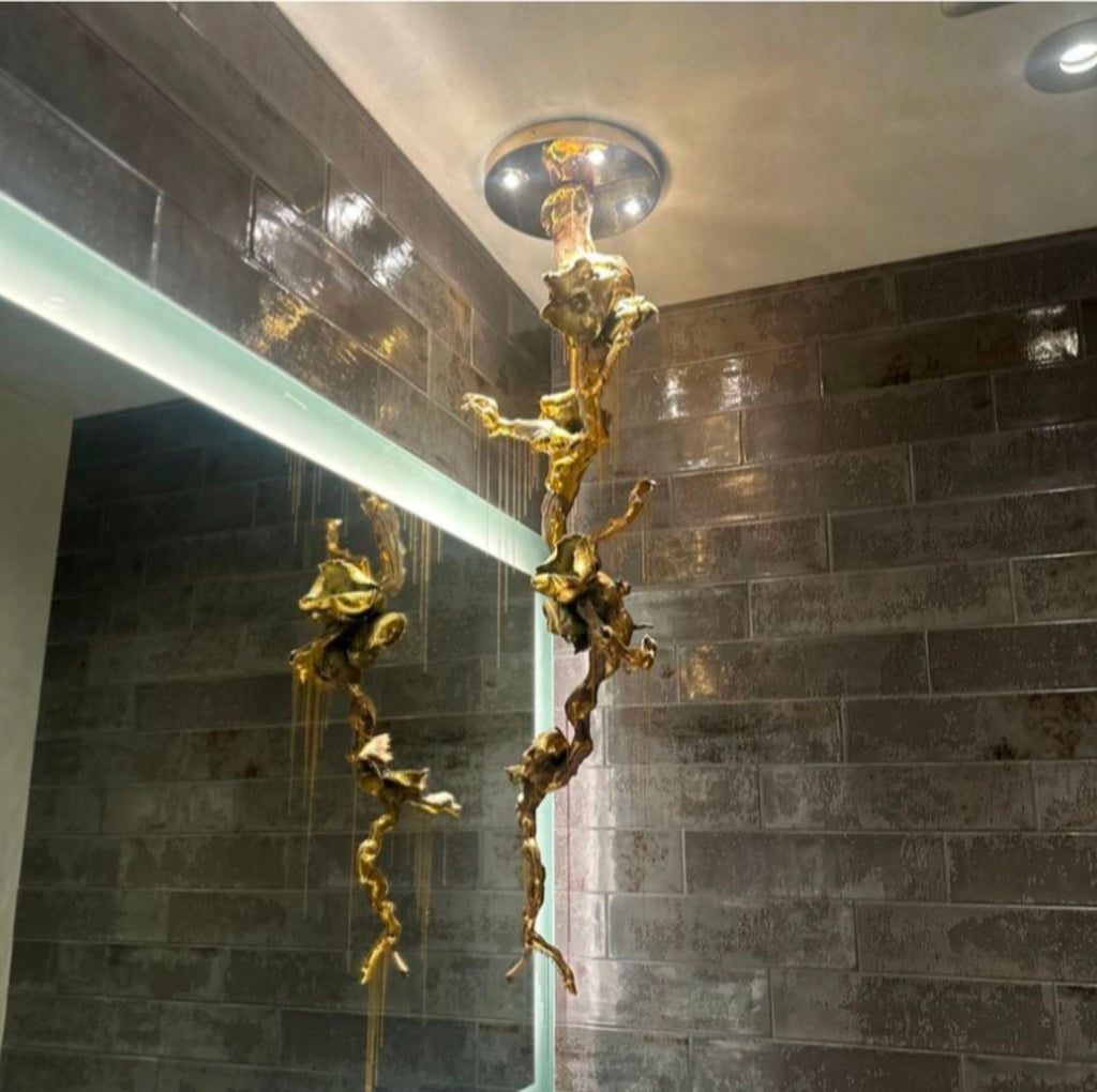 Sculptural gold ceiling light fixture by Jocelyn Marsh, reflected in a bathroom mirror with dark tiled walls.