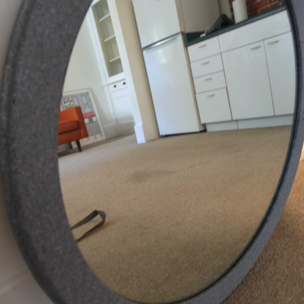 1. Round mirror with a textured dark frame reflecting a kitchen and living room area with beige carpet.  