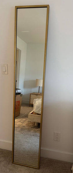 1. Tall rectangular West Elm mirror with a gold frame, leaning against a white wall in a bedroom setting.