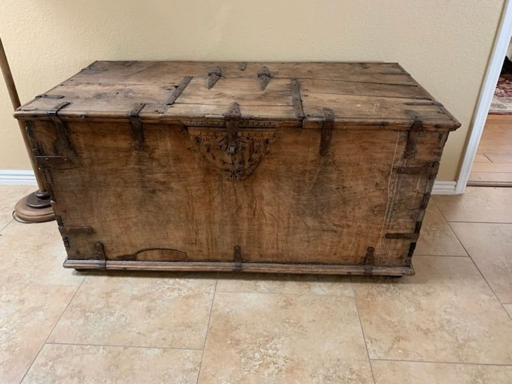 A large, rustic wooden trunk by Reperch with metal hinges and decorative detailing sits on a tiled floor.