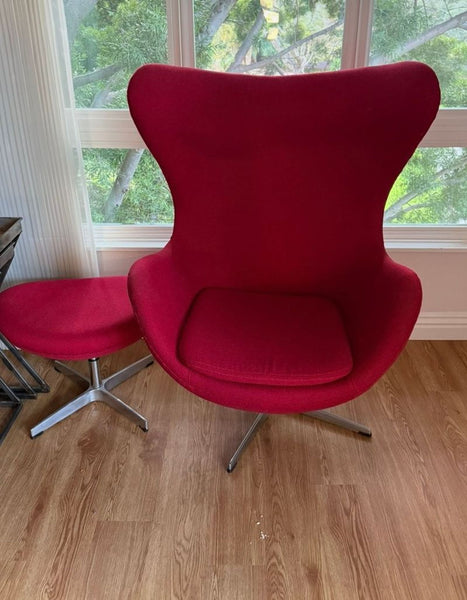 Modern red lounge chair with matching ottoman on metal legs, placed near a window with wooden flooring.