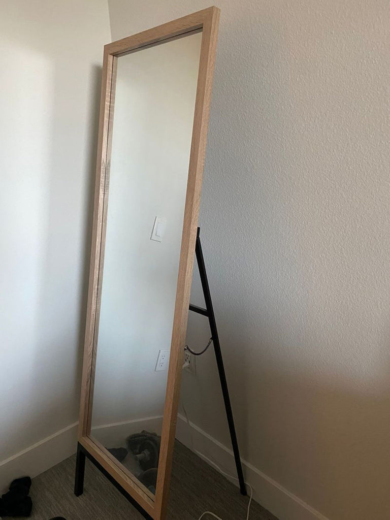 1. Full-length rectangular mirror with a light wood frame and black stand, placed in a corner against white walls.