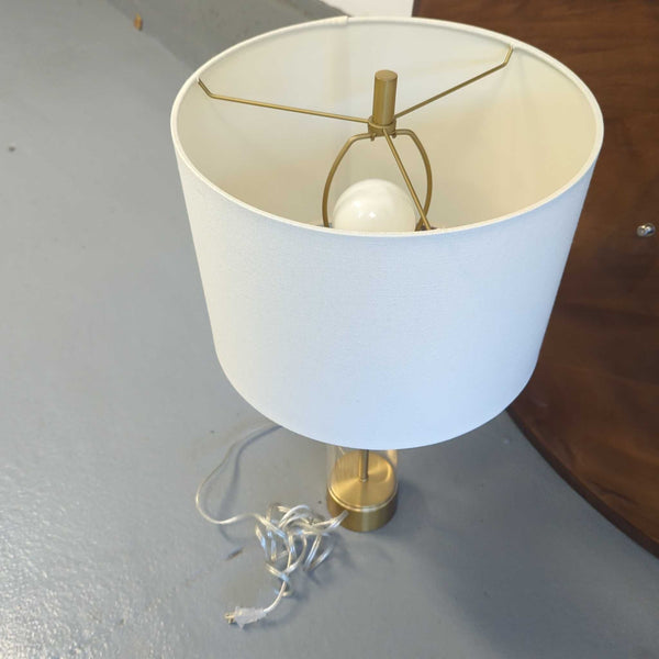 1. Modern table lamp with a white fabric shade, clear cylindrical base, and gold accents, on a gray floor.