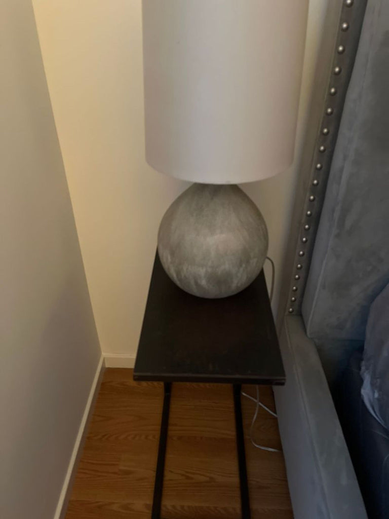 1. Grey textured table lamp with a round base and cream shade placed on a small black side table by a bed.