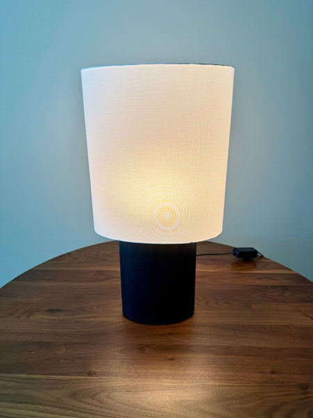 1. Room & Board table lamp with a cylindrical black base and a white drum shade, turned on, on a wood table.