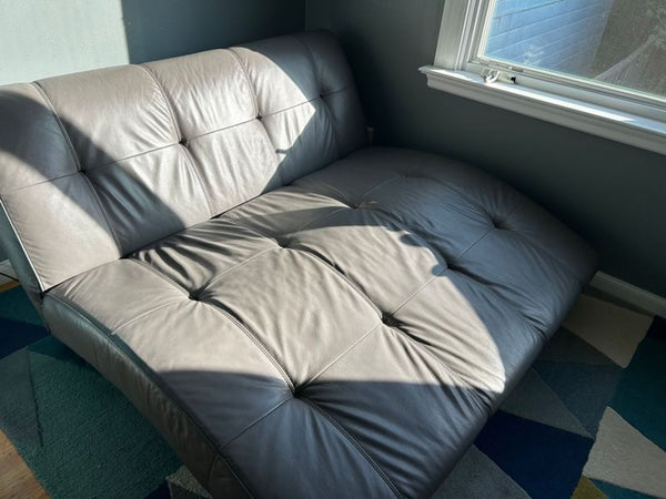 1. Gray tufted EQ3 futon with leather upholstery, positioned by a window on a patterned rug in sunlight.  