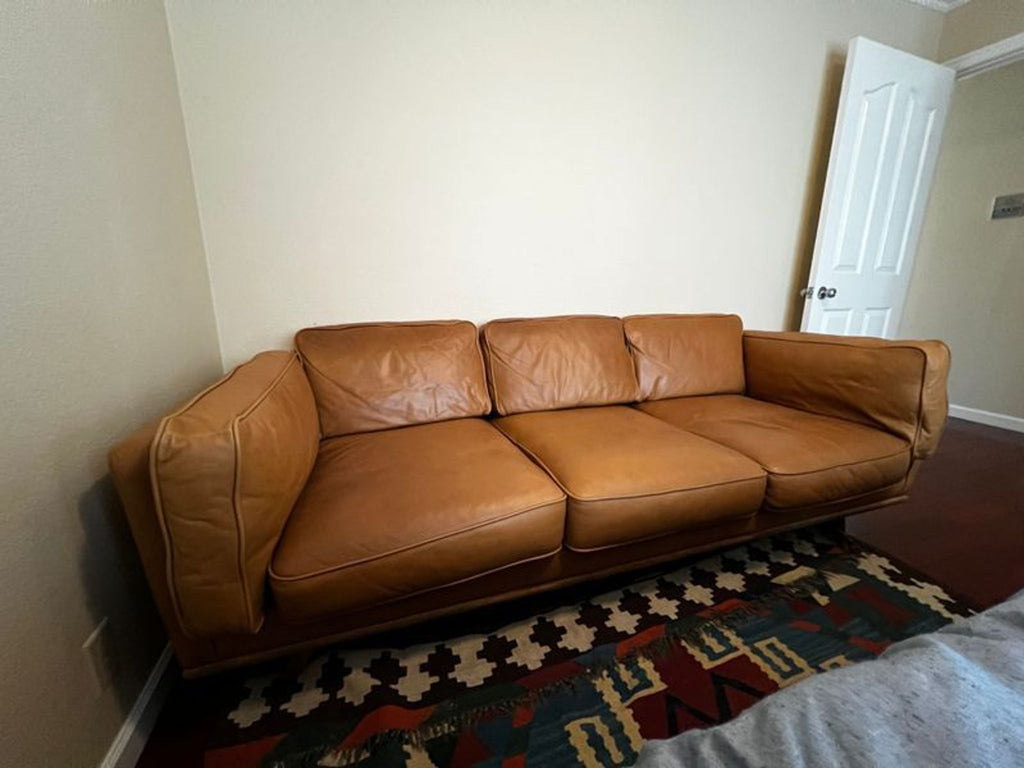 Tan Article couch in a small room, positioned on a colorful rug near a bed.