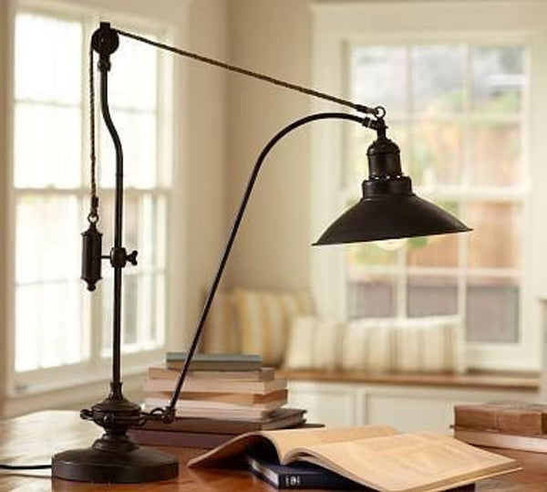 1. "Pottery Barn industrial-style lamp with pulley system on a desk, books scattered around, with a warm room backdrop."