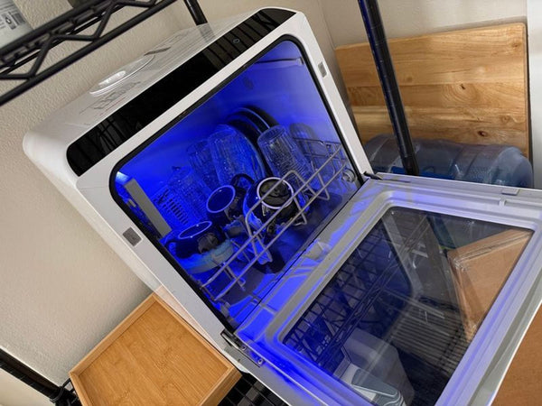 1. Compact countertop dishwasher with glass door open, blue interior light, and dishes loaded inside for washing.