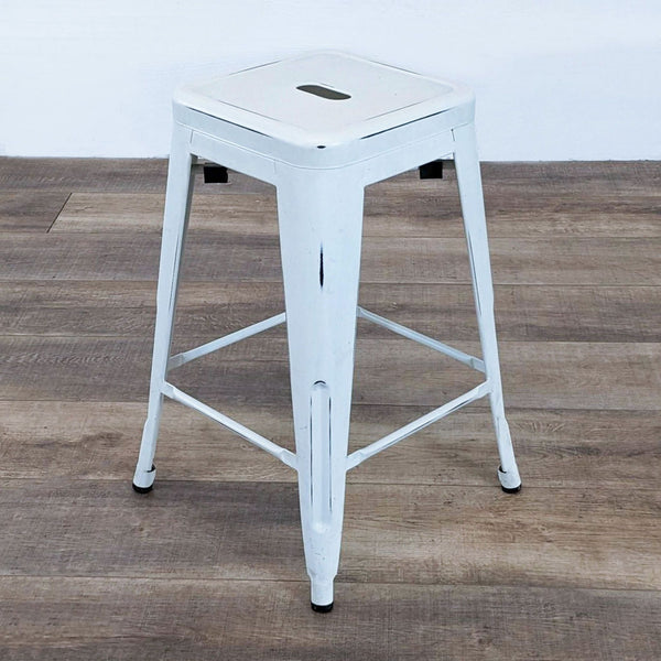 White distressed metal stool by Reperch with a square seat and footrest on a wooden floor.