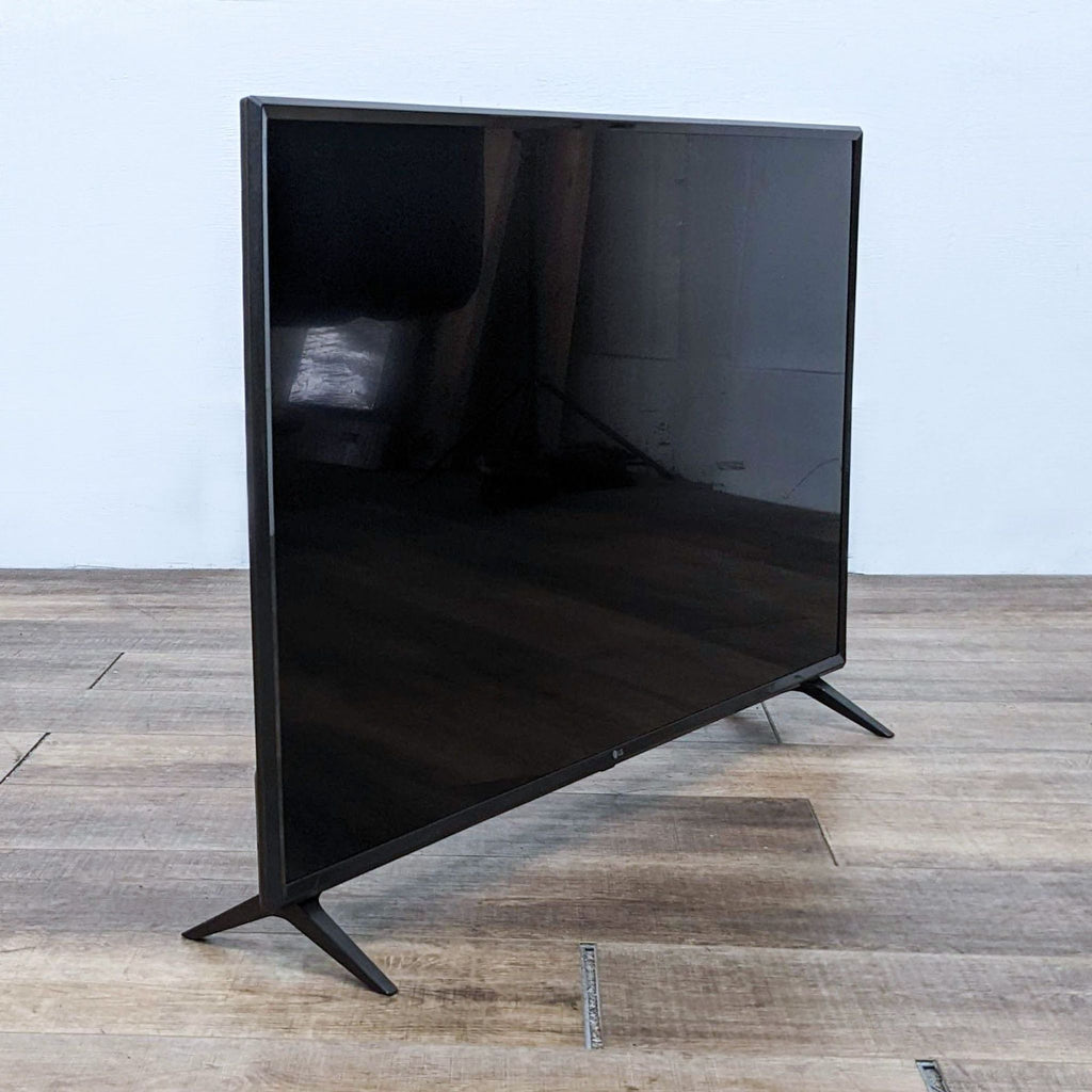 LG TV turned off, showing a reflective black screen with sturdy legs, placed on wooden flooring against a bare wall.