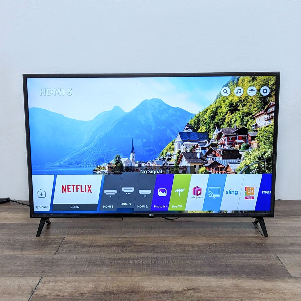 LG TV displaying colorful menu with scenic background on home screen, standing on a wooden floor against a white wall.