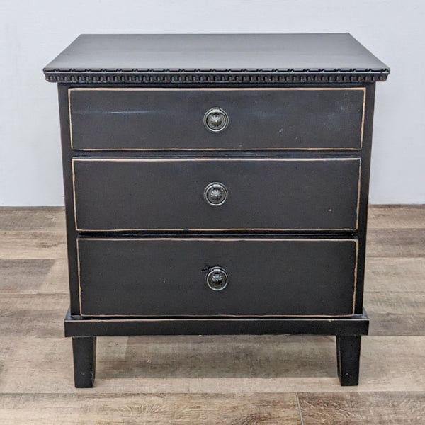 Black Harvest end table with decorative moulding and three drawers with pull-ring handles, against a wood floor.