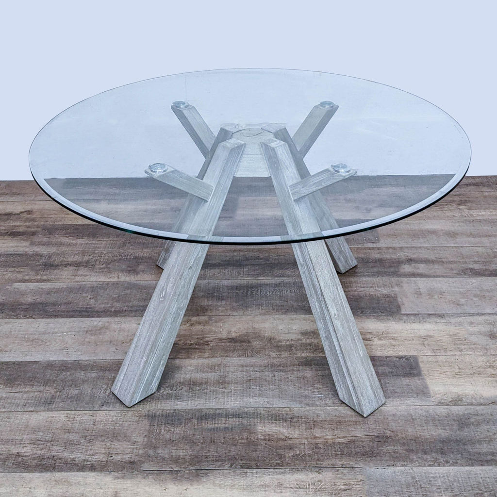 Round glass dining table by Reperch, featuring bleached acacia wood legs in flaired design, positioned on plank-style flooring.