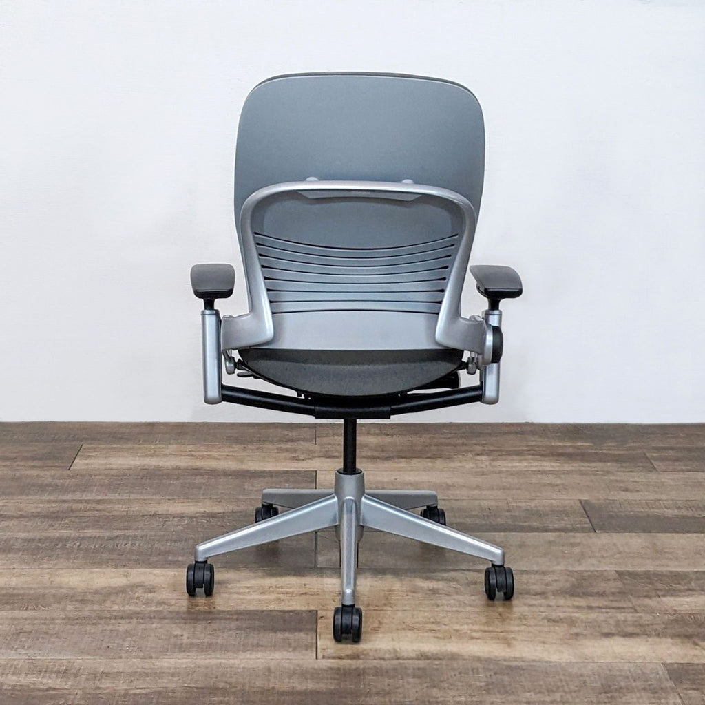 Gray Steelcase Store office chair with a high back and lumbar support, viewed from behind on a wooden floor with a white wall.