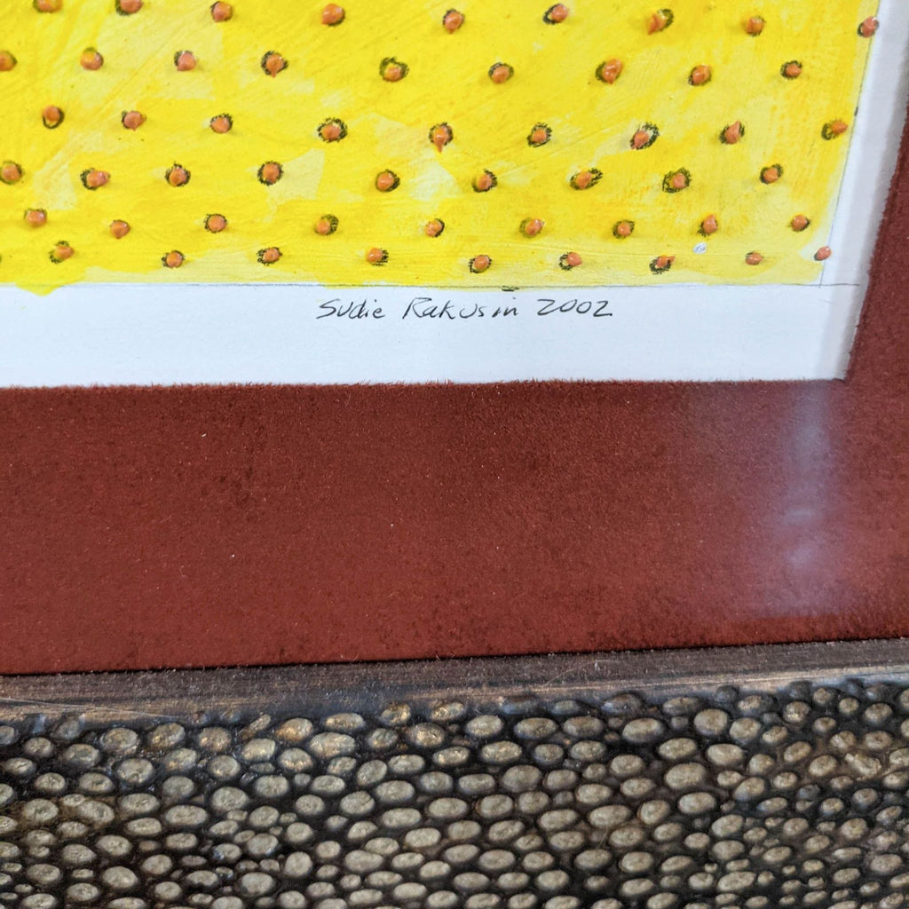 2. Close-up of signature "Sudie Rakusin 2002" on a yellow-patterned section of the framed art print.